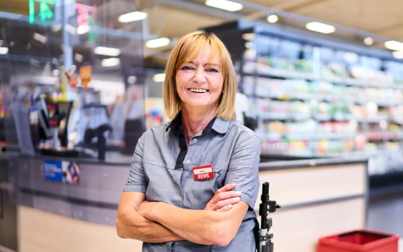 Eine Frau im Rollstuhl, traegt Rewe Arbeitskleidung und befindet sich in einer Rewe Filiale.