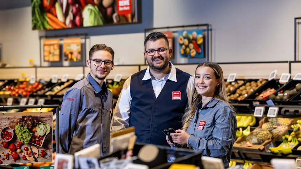 Drei REWE Mitarbeitende stehen gemeinsam in der Obstabteilung eines REWE Markts.