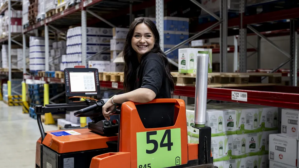 Eine REWE Auszubildende zur Fachkraft für Lagerlogistik faehrt einen Transportwagen im Lager und lacht